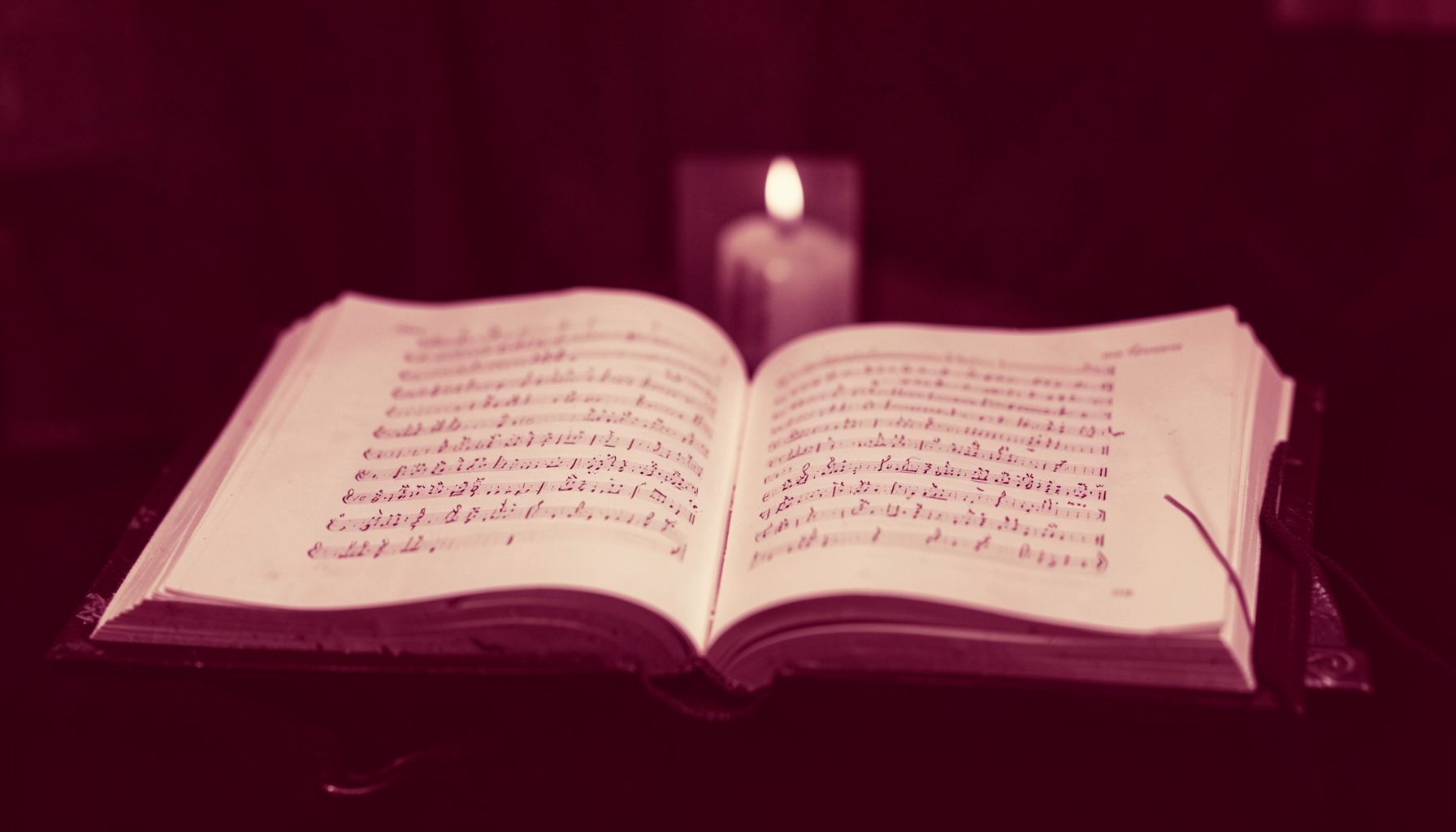 A duotone photograph in deep burgundy and cream tones showing an open liturgical choir book resting on a wooden stand, with soft candlelight illuminating the chant notation.
